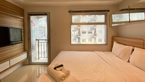 A hotel room with a bed, a TV mounted on the wall, a window with a view of buildings, and a door leading to an outdoor balcony. - Artsy Private Studio Room At Grand Asia Afrika Bandung Apartment (Bandung)