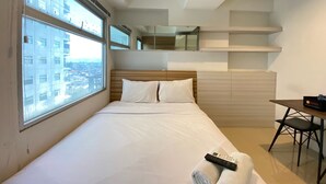 A modern hotel room with a large bed, a desk, and a chair. There is a window with a city view, and a mirror above the bed. - Artsy Private Studio Room At Grand Asia Afrika Bandung Apartment (Bandung)