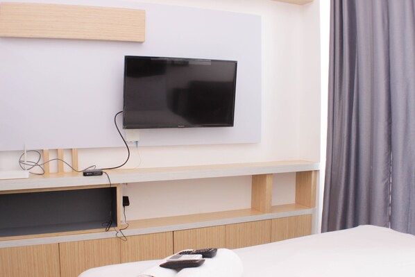 Une chambre d’hôtel avec un téléviseur fixé au mur, un lit avec une tête de lit blanche et un meuble en bois placé sous le téléviseur.