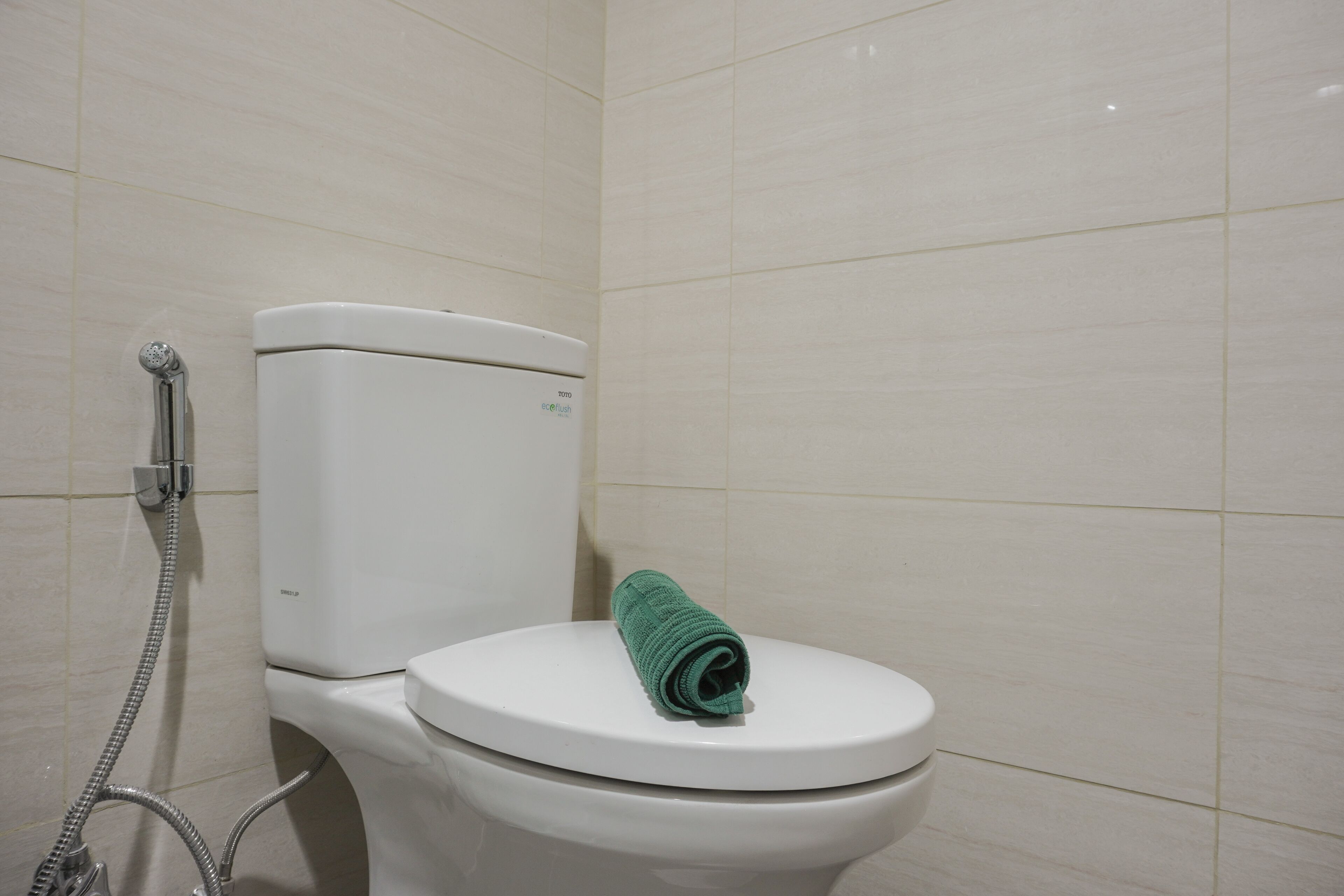 Un inodoro blanco con una toalla verde sobre el asiento, un cabezal de ducha y una pared con azulejos de colores claros.