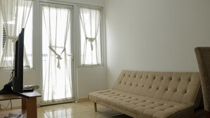 A room with a beige sofa, a TV on a wooden table, and a window with sheer curtains. - Spacious And Elegant 3Br At Grand Palace Kemayoran Apartment (Jakarta)