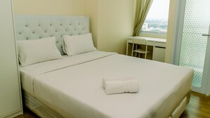 A neatly made bed with white linens and a folded towel, a tufted headboard, and a window with curtains. - Warm And Cozy Studio Room Apartment At B Residence (Pagedangan)
