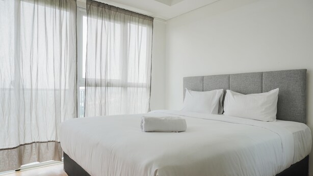 A neatly made bed with a grey headboard and white bedding in a bright room with large windows and sheer curtains. - Nice And Cozy Studio Room At Casa De Parco Apartment (Pagedangan)