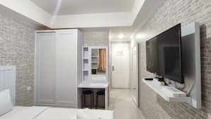 A modern hotel room with a bed, a TV mounted on the wall, a wardrobe, and a small seating area with stools.