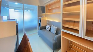 A modern room with a grey sofa, a wooden chair, and a large white shelving unit. - Minimalist And Nice 1Br At Gateway Pasteur Apartment (Cimahi)