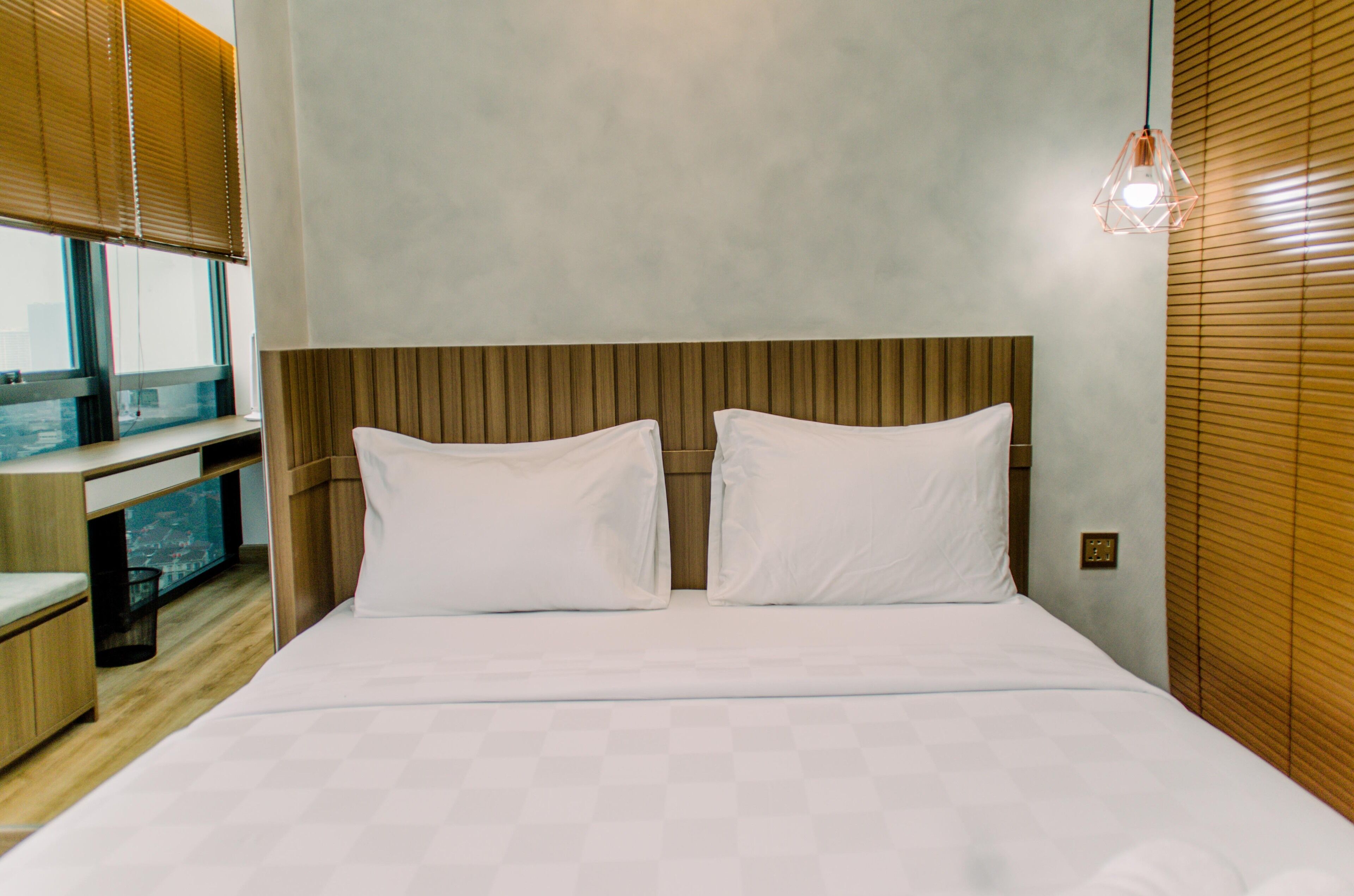 Hotelkamer met een houten hoofdbord, een bed met wit bedgoed, een klein bureau met stoel en een raam met zonwering.