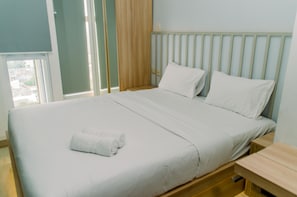A neatly made bed with white linens and a rolled towel, a wooden headboard, and a wooden nightstand with a drawer.