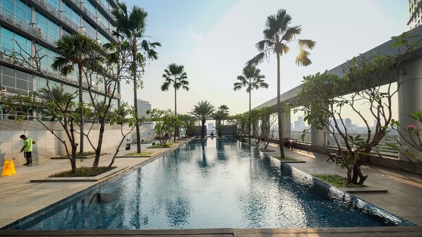 A swimming pool with palm trees, a person cleaning, and modern buildings in the background. - Stunning And Comfy Studio Apartment At Citylofts Sudirman (Jakarta)