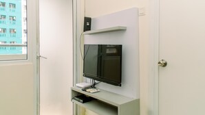 A room with a mounted flat-screen TV on a white wall, a small shelf with a DVD player, and a window with a view of a blue building. - Modern Look 2Br Green Pramuka City Apartment Near Mall (Jakarta)