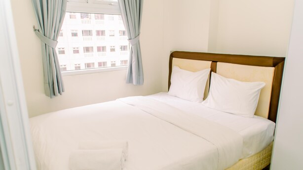 A neatly made bed with white linens and pillows, a wooden headboard, and a window with curtains. - Modern Look 2Br Green Pramuka City Apartment Near Mall (Jakarta)