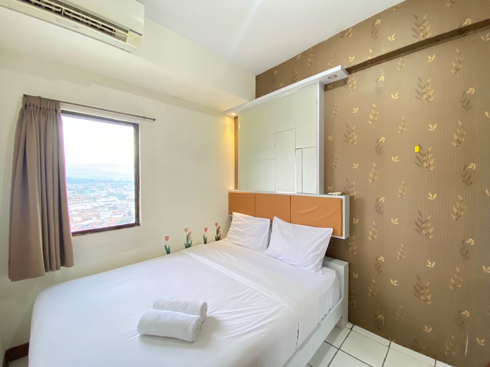 A hotel room with a large bed, a window with curtains, a wall-mounted air conditioner, and a view of the city.