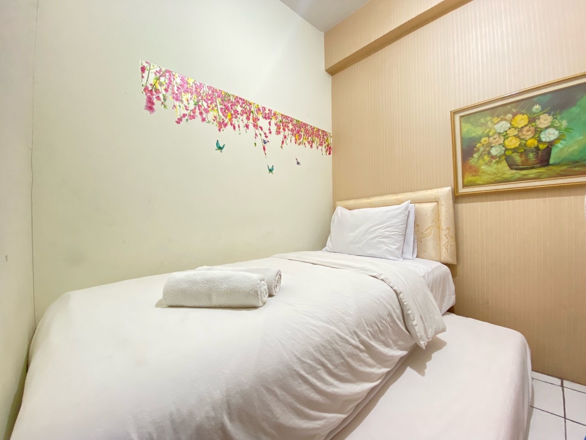 A neatly made bed with white linens and a rolled towel, a floral wall decal, and a framed painting of flowers on the wall.