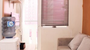 A living room with a sofa, a water dispenser, and a window with blinds. - Comfort 2Br At Green Pramuka Apartment (Jakarta)