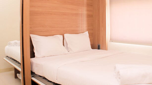 A neatly made bed with white linens and pillows, a wooden headboard, and a small shelf with towels. - Comfort 2Br At Green Pramuka Apartment (Jakarta)