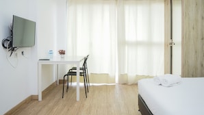 A hotel room with a bed, a desk, a chair, a television, and a window with curtains. - Fancy And Nice Studio Room At Casa De Parco Apartment (Pagedangan)