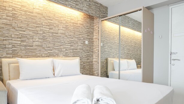 A modern hotel room with a stone wall, a large bed with white linens, and a mirrored closet. - Well Designed And Cozy Studio At Bassura City Apartment (Jakarta)