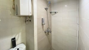 Un baño pequeño y limpio con azulejos blancos, inodoro, ducha con cabezal de ducha manual y un calentador de pared.