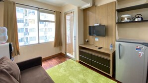 Un espacio habitable compacto con sofá, alfombra verde, televisor, refrigerador y estante de madera con utensilios de cocina.