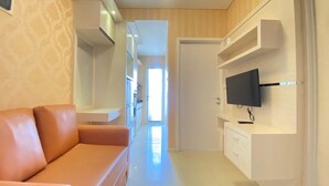 A modern living room with a sofa, a flat-screen TV mounted on a wall, and a kitchen area in the background. - Classic Private 1Br Apartment At Parahyangan Residence Bandung (Bandung)