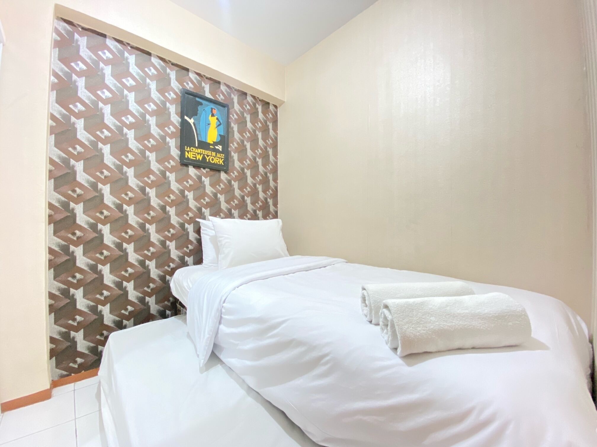 Una cama bien hecha con ropa blanca y una toalla doblada, una pared con estampado geométrico y un cartel de un músico de jazz.