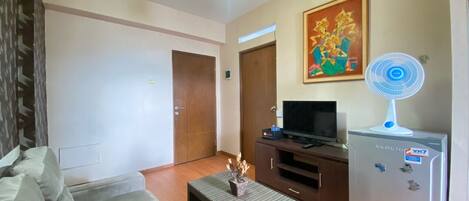 A living room with a sofa, a small table, a TV, a refrigerator, and a fan.