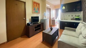 A living room with a sofa, a TV on a wooden stand, a small table with a plant, a kitchen area with cabinets and a microwave, and a door with a number. - Spacious 2Br At Gateway Ahmad Yani Apartment (Bandung)