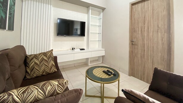 A modern living room with a sofa, a coffee table, a TV mounted on a white wall, and a wooden door. - Cozy 2Br Apartment At Newton Residence Bandung (Bandung)