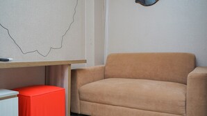 Um quarto com um sofá bege, um banquinho vermelho e uma escrivaninha branca com uma caneta preta.