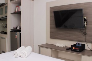 A hotel room with a bed, a TV mounted on the wall, a small desk with a phone and a box of toiletries. - Cozy And Warm Studio At Grand Sentraland Karawang Apartment (Karawang)