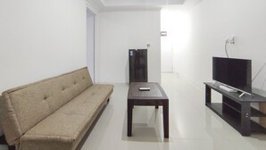 A minimalist living room with a beige sofa, a dark wooden coffee table, a television on a stand, and a flat-screen TV on a black stand. - Good Deal 1Br At Paltrow City Apartment (Semarang)