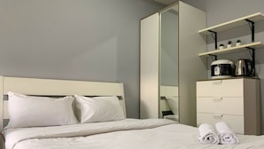 A neatly made bed with white linens and pillows, a tall wardrobe with a mirror, and a shelf with a kettle and a coffee maker. - Tranquil Living Studio At Belmont Residence Puri Apartment (Jakarta)