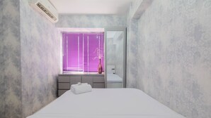 A modern bedroom with a large bed, a window with purple blinds, a bedside table with a lamp, and a mirror. - Strategic And Restful 2Br At Bassura City Apartment (Jakarta)