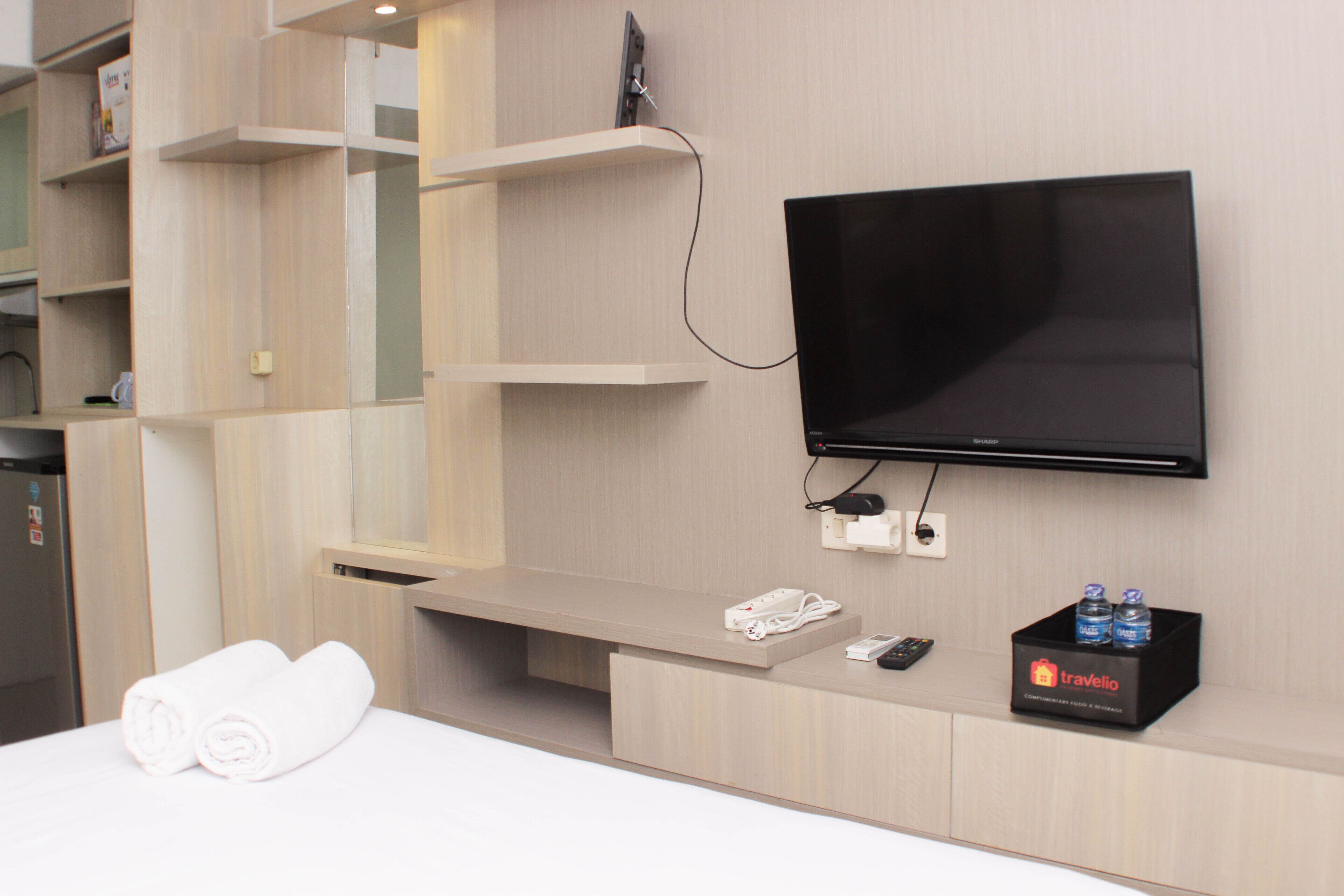 Habitación de hotel con televisión de pantalla plana montada en la pared, una mesita de noche con un control remoto, un enchufe y una mesita auxiliar.