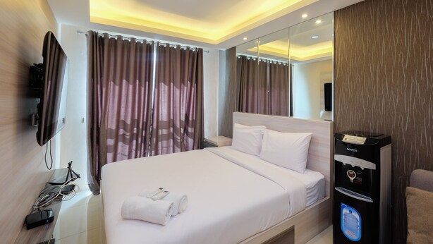 A modern hotel room with a large bed, a flat-screen TV mounted on the wall, a built-in water dispenser, and a mirror above the bed. - Modern And Comfy 1Br Apartment At Woodland Park Residence (Jakarta)