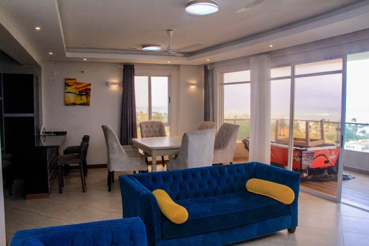 A spacious living area with a dining table, multiple chairs, a blue sofa, and large windows offering a view of the outdoors.