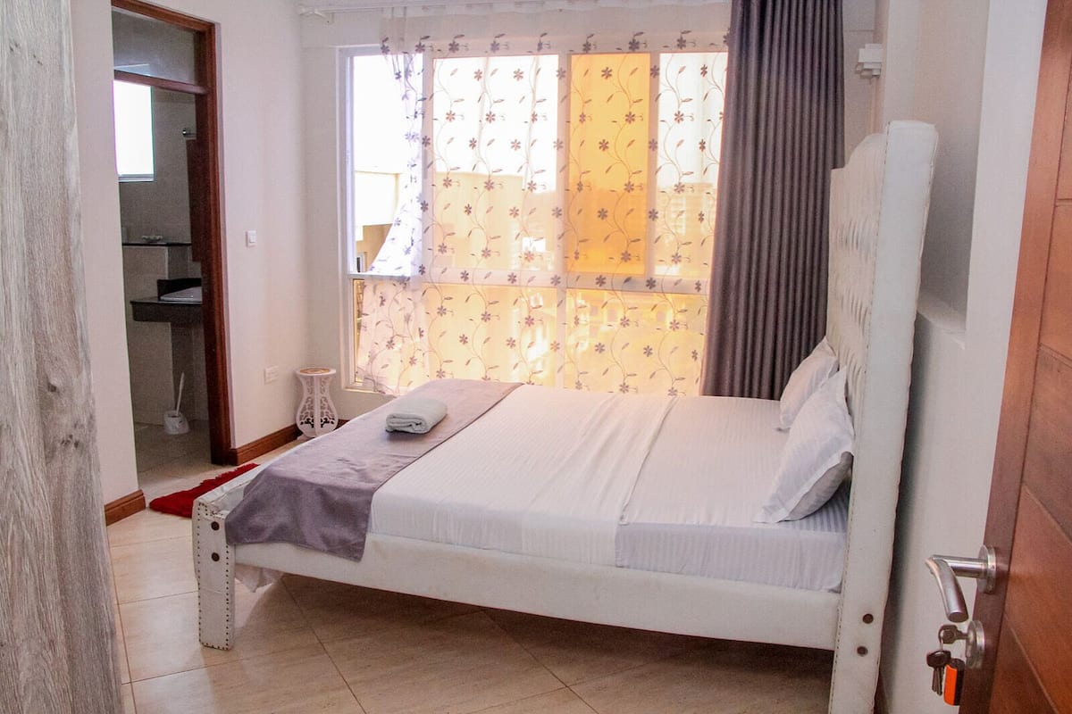 A bedroom with a bed, a window with floral curtains, a bedside table, and a door leading to a bathroom.