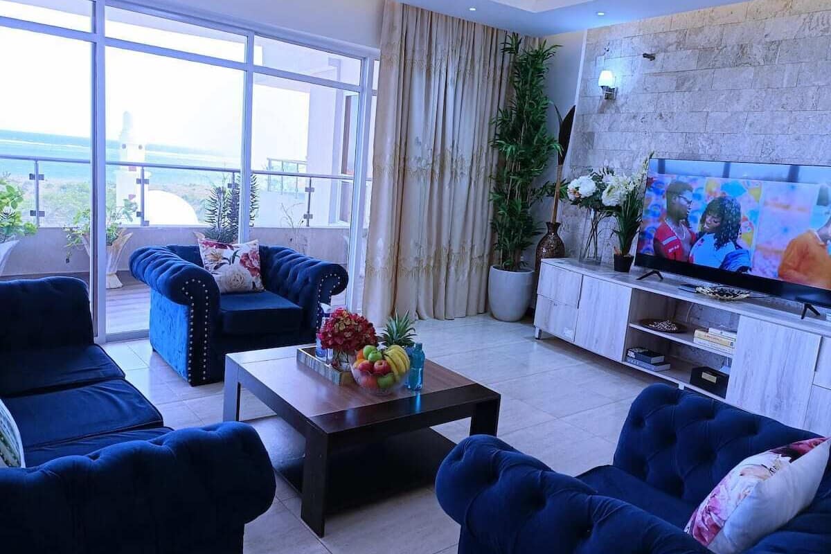 A modern living room with blue sofas, a coffee table with fruit, a TV, and a view of the outdoors.