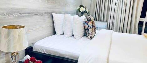 A hotel room with a bed, a nightstand with a lamp, a vase with flowers, and a view of the outside through curtains.
