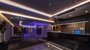 A modern hotel reception area with a dark reception desk, a digital screen, and a staircase leading to upper floors. - HOTEL ZEN RIKYU CHIBA-SHIROI (Shiroi)