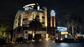 A modern hotel with a prominent 'HOTEL WITH ZEN' sign, a well-lit entrance, and a palm tree in the foreground. - HOTEL ZEN RIKYU CHIBA-SHIROI (Shiroi)