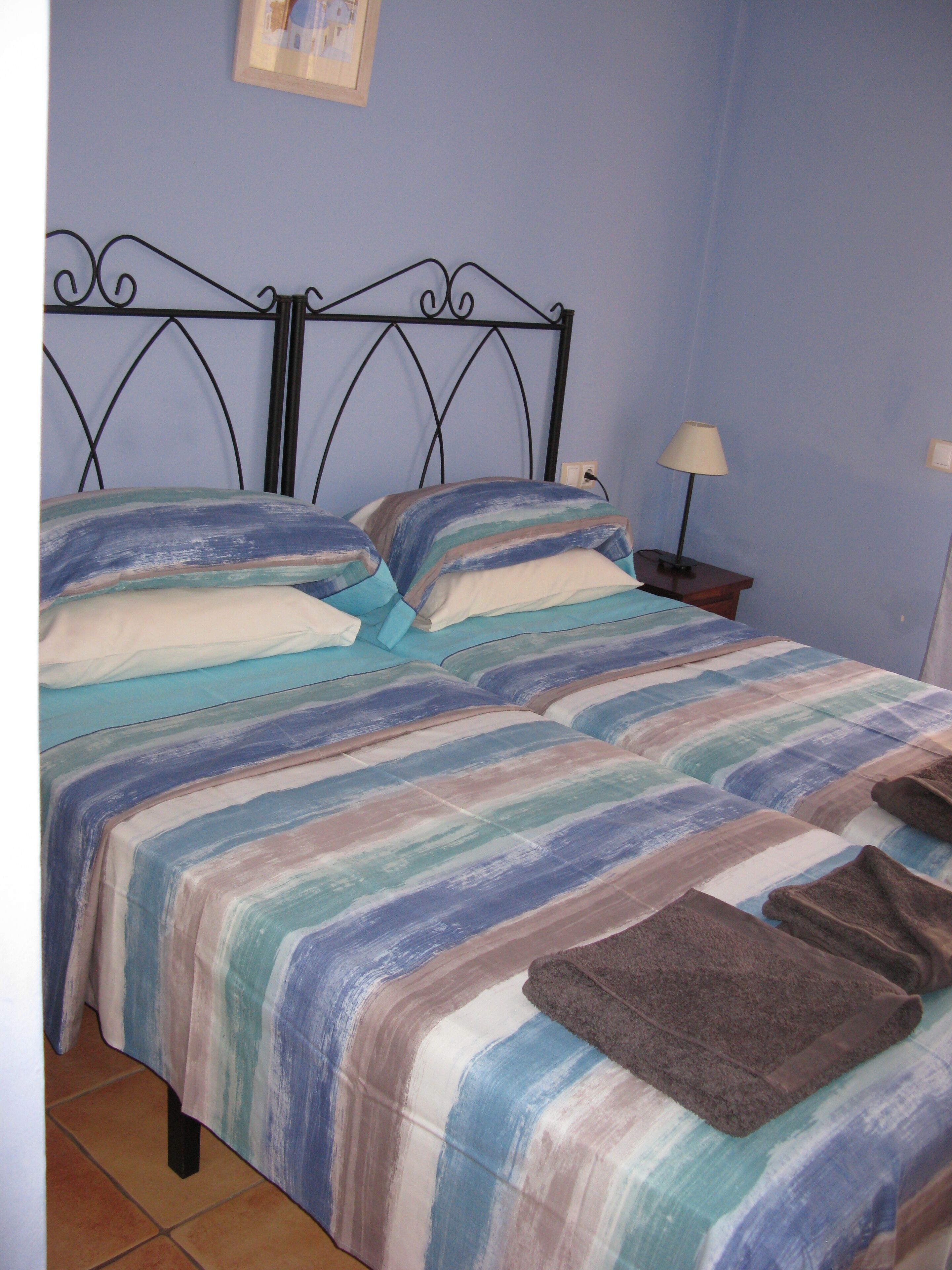 A bed with a striped bedspread and pillows, a bedside table with a lamp, and a wall-mounted power outlet.