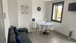Una sala de estar moderna con un sofá gris, una mesa de comedor redonda con sillas blancas, una mesita blanca con un microondas, un reloj en la pared y un televisor montado.