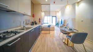 A modern kitchen with a dining area, wooden flooring, and a large window providing natural light. - JVC Studio park view fully equipped - 360 vacation (Dubai)