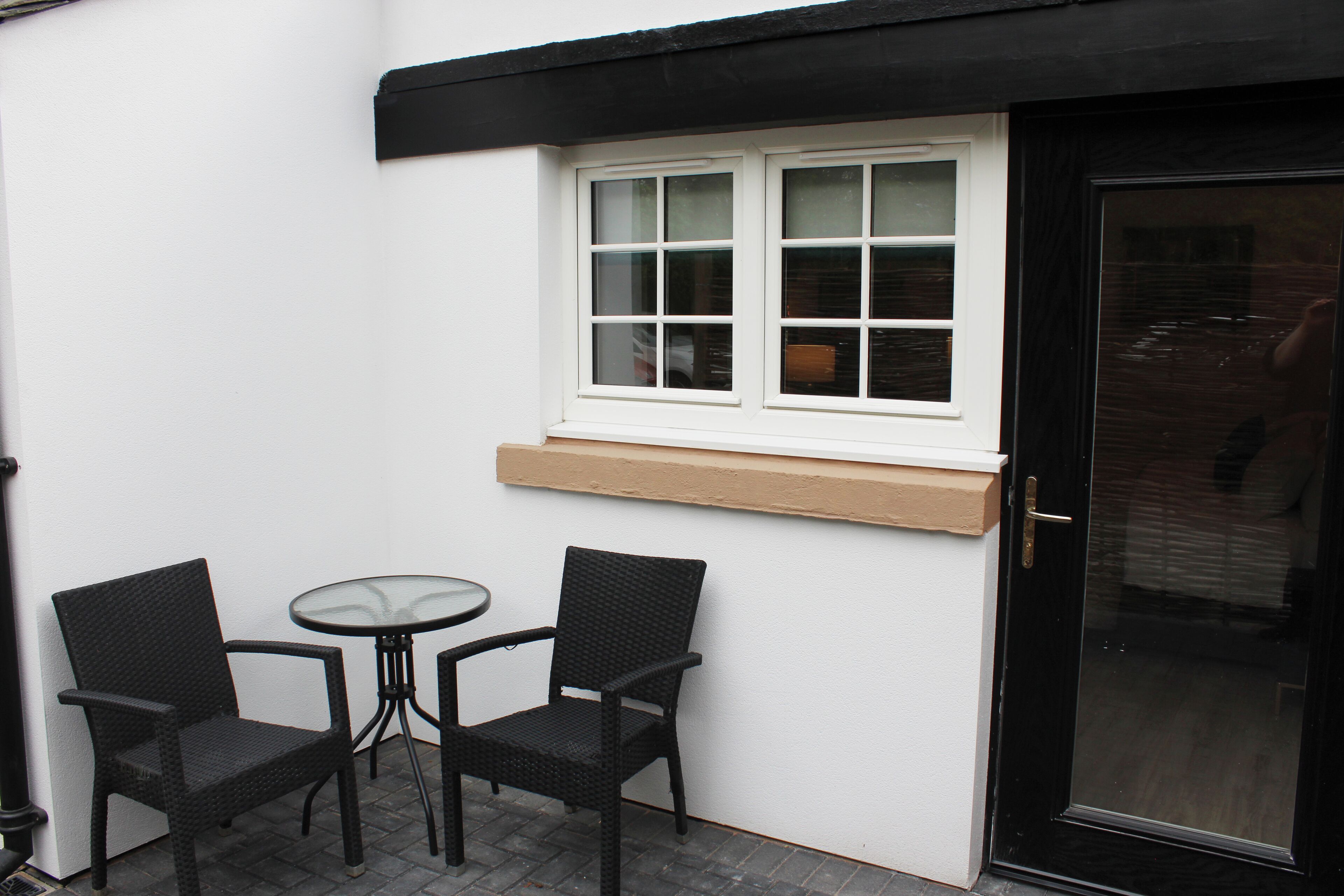 A small outdoor seating area with two wicker chairs and a glass table, adjacent to a white wall with a window and a black door.