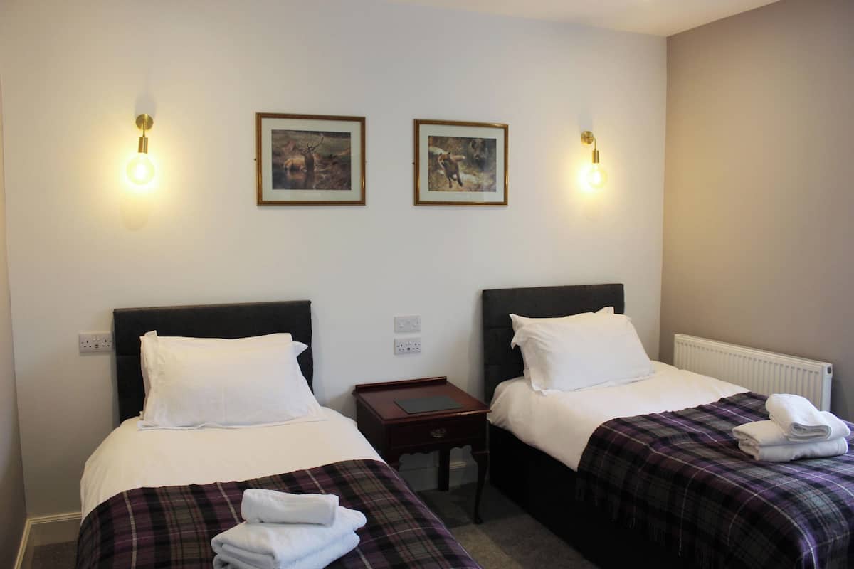 Ein Hotelzimmer mit zwei Betten, jeweils mit weißen Bettwäsche und einer karierten Decke. An den Wänden sind Wandleuchten und zwei gerahmte Bilder angebracht.