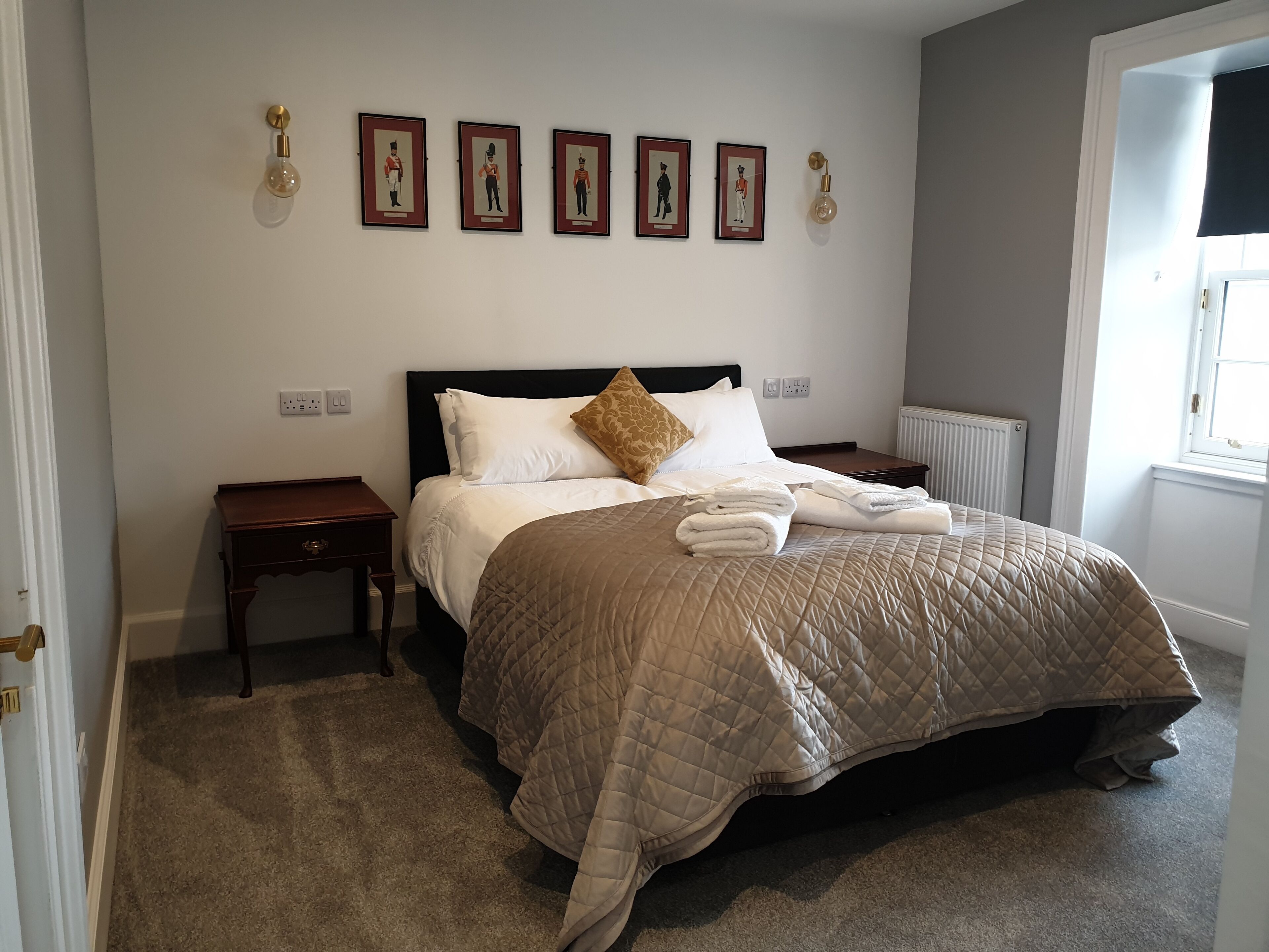 Un lit bien fait, recouvert d’une courtepointe matelassée, une table de chevet en bois et un luminaire fixé au mur.