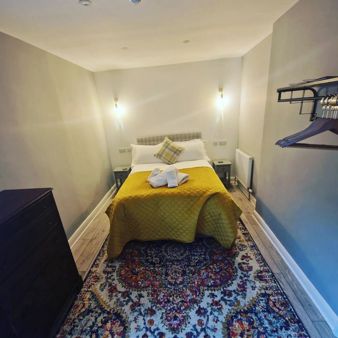 A hotel room with a bed, a nightstand, a dresser, and a wall-mounted coat rack.
