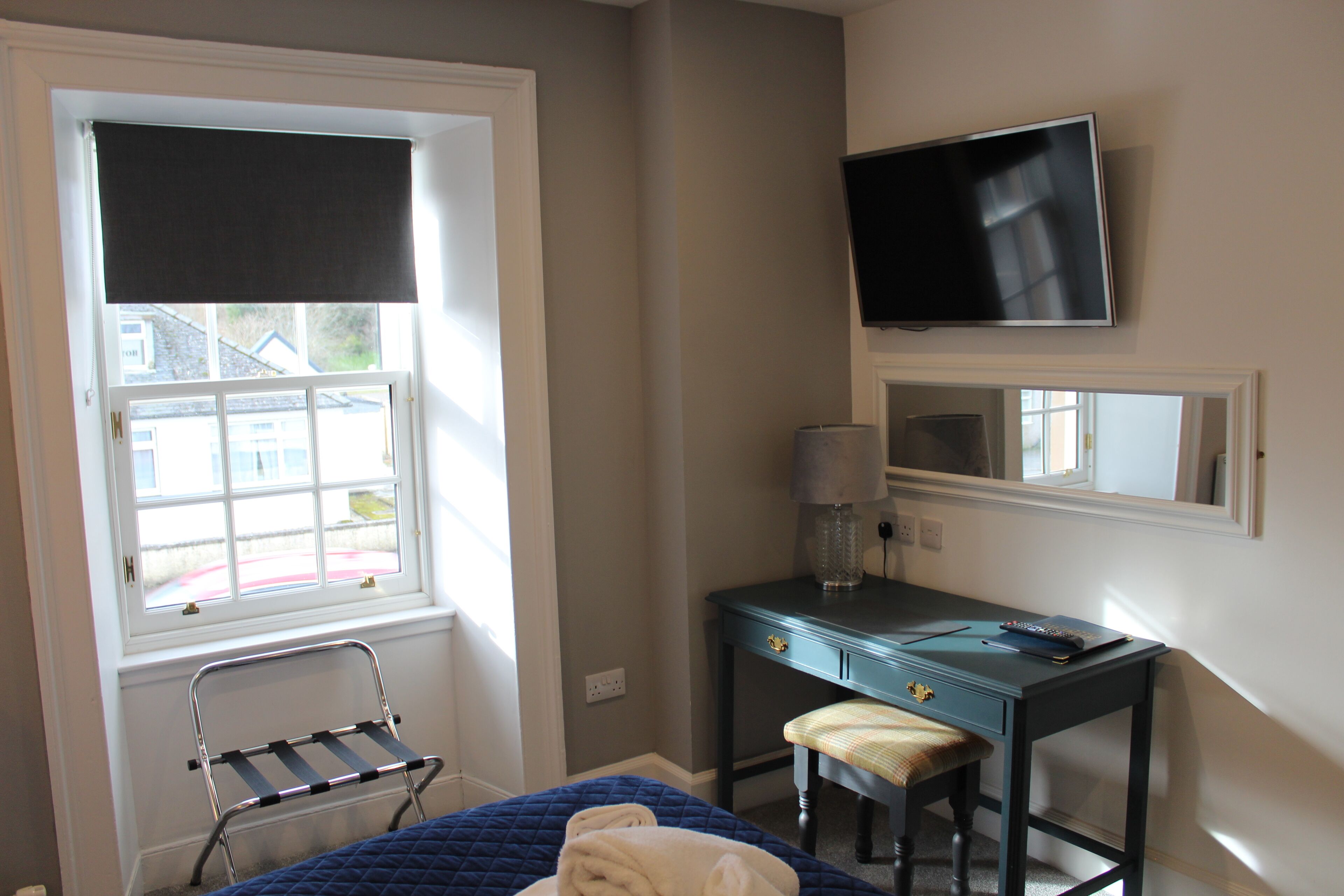 A bedroom with a bed, a desk, a chair, a TV, and a window with a view of houses.