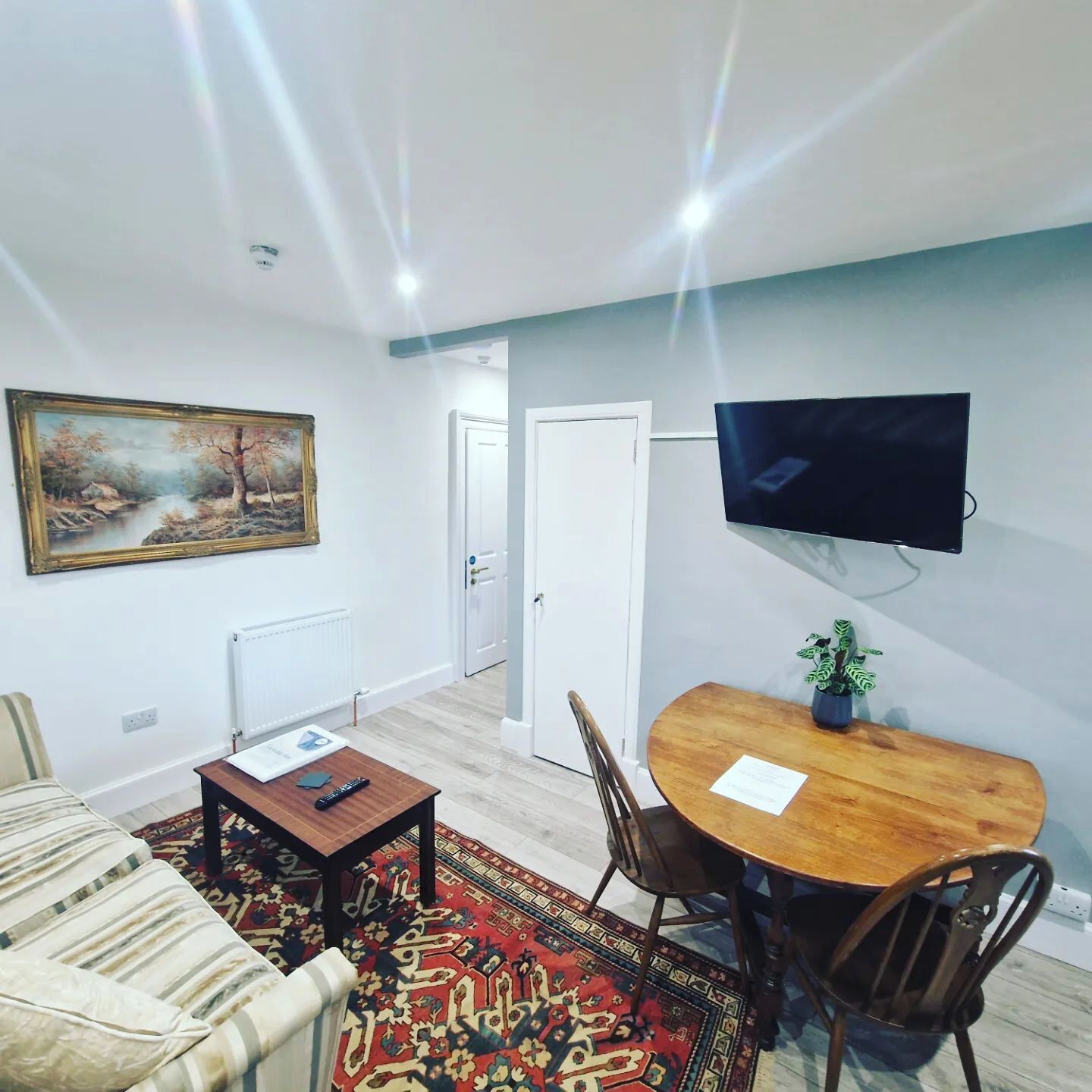 A living room with a sofa, a coffee table, a TV mounted on the wall, a dining table with chairs, and a painting on the wall.