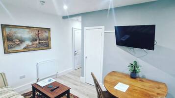 A living room with a sofa, a coffee table, a TV mounted on the wall, a dining table with chairs, and a painting on the wall.